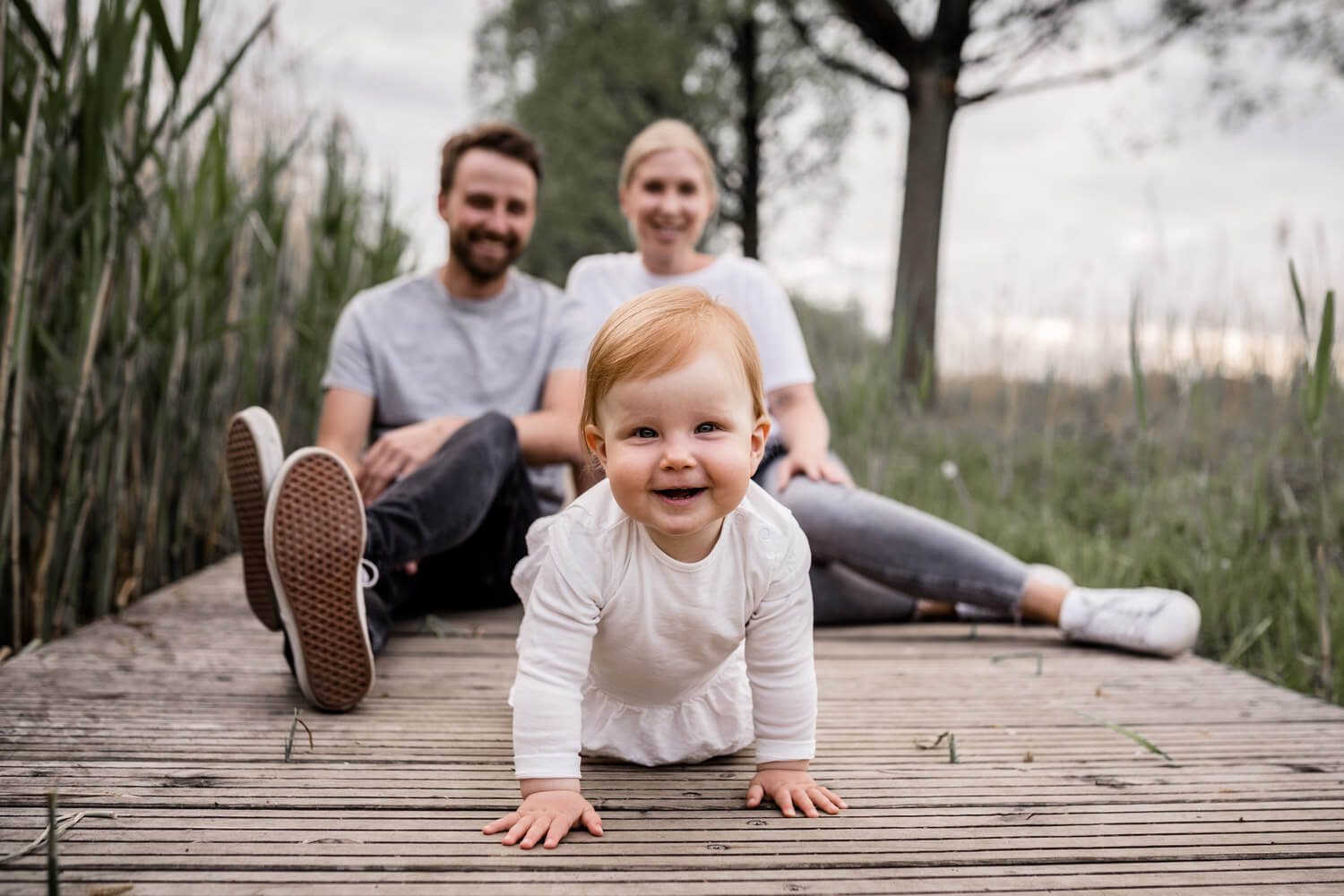 Kleines Mädchen krabbelt auf Holzsteg, lacht in Kamera, Eltern sitzen im Hintergrund, Familienbilder in Isernhagen