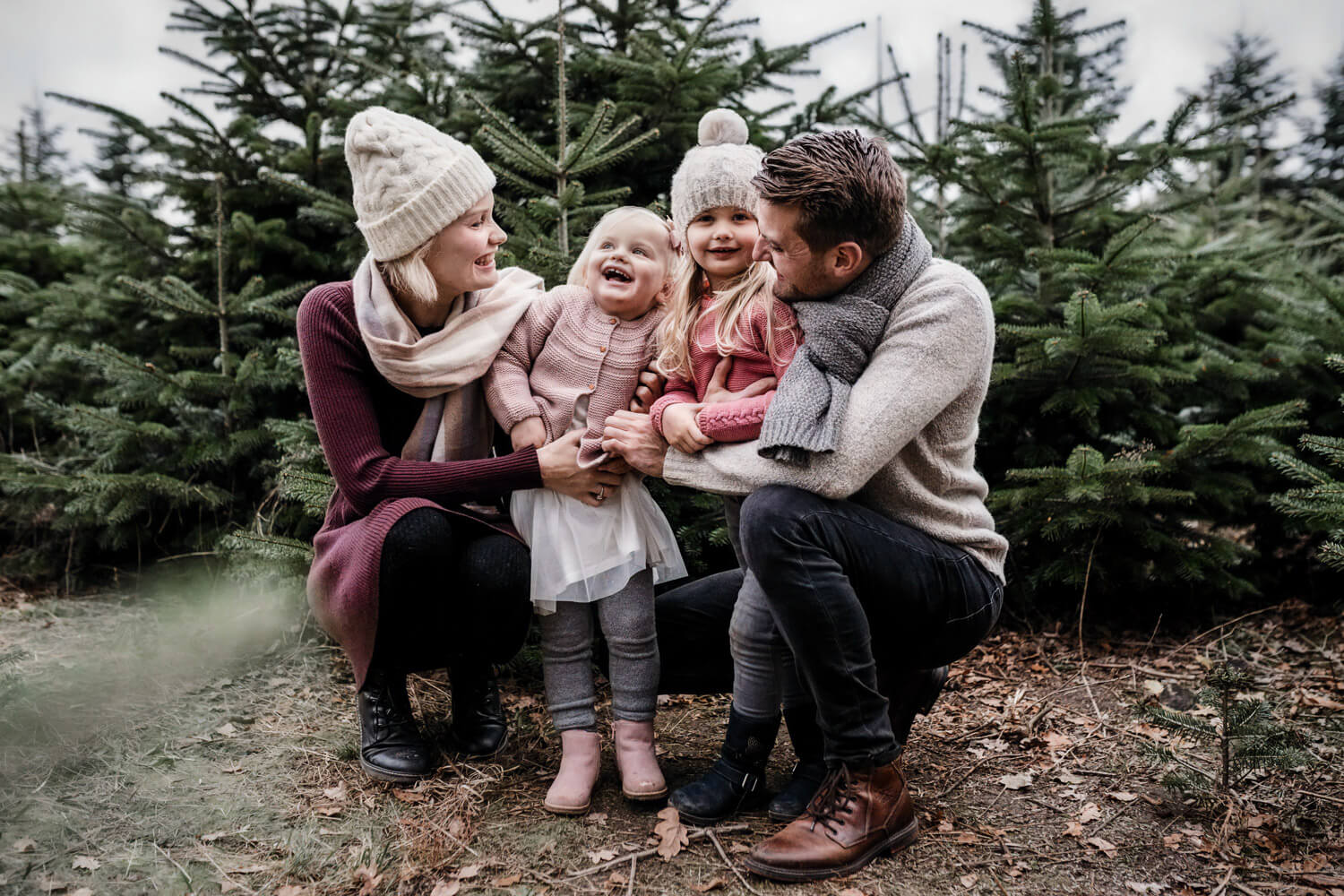 Vierköpfige Familie hockt draußen zwischen kleinen Weihnachtsbäumen, zwei Töchter stehen zwischen ihren Eltern, lachen