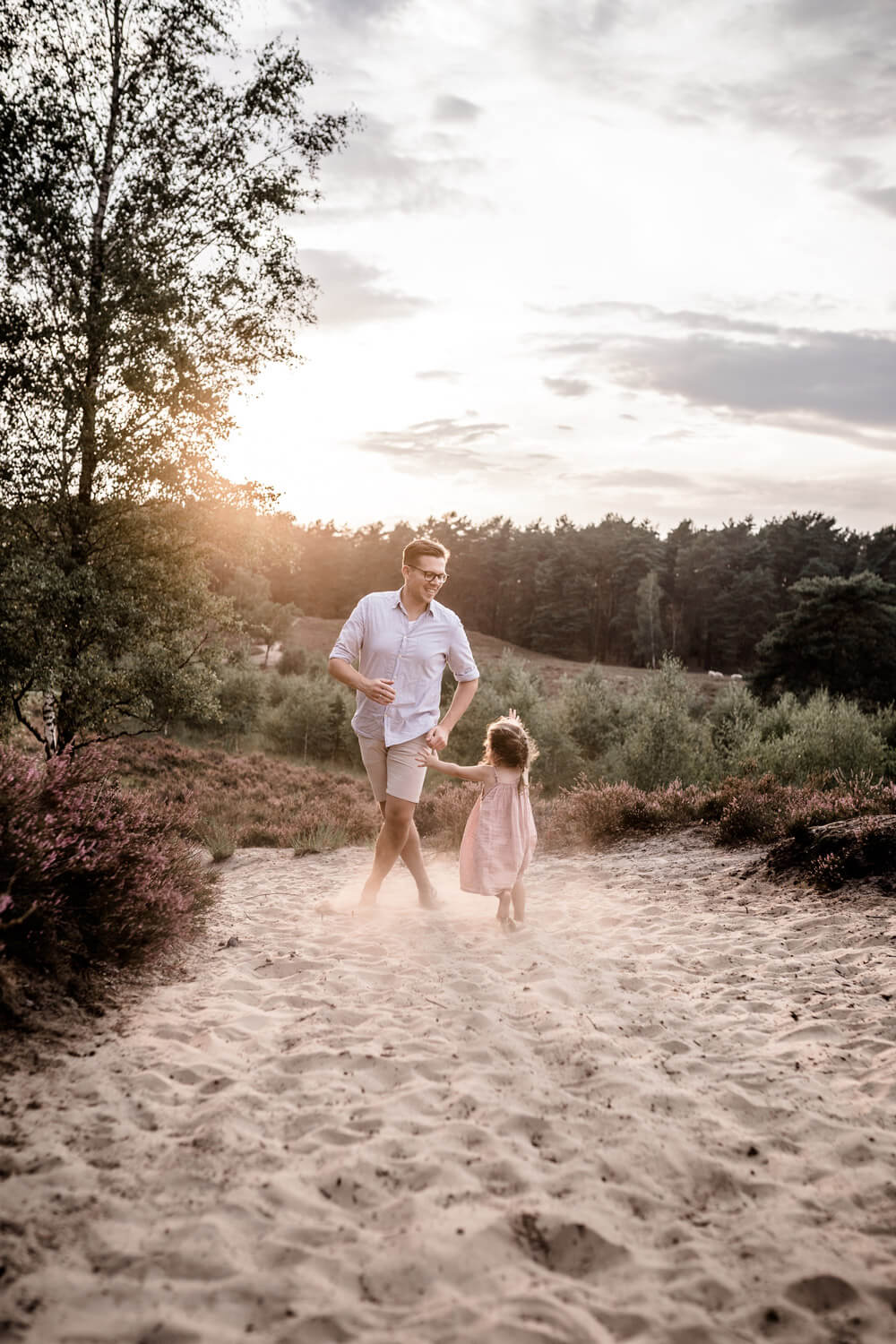 Vater spielt mit Tochter fangen im Sand in Heide, Mädchen trägt Kleid, Sonnenuntergang während Familienshooting