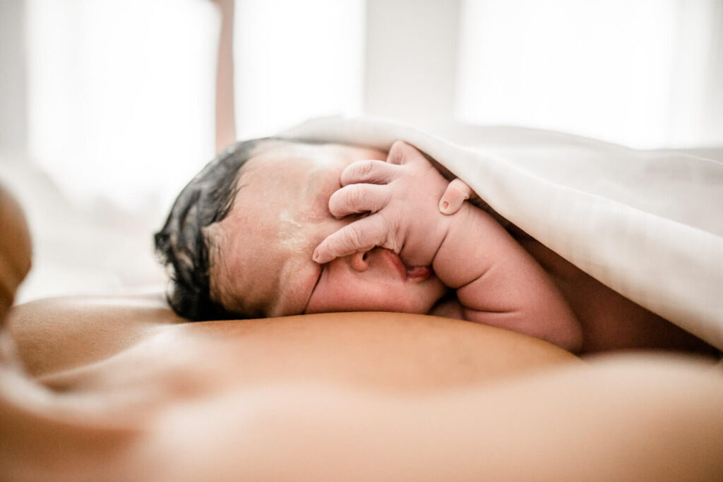 Gerade geborenens Baby liegt auf Brust des Vater, nuckelt an Daumen, in Handtuch gewickelt