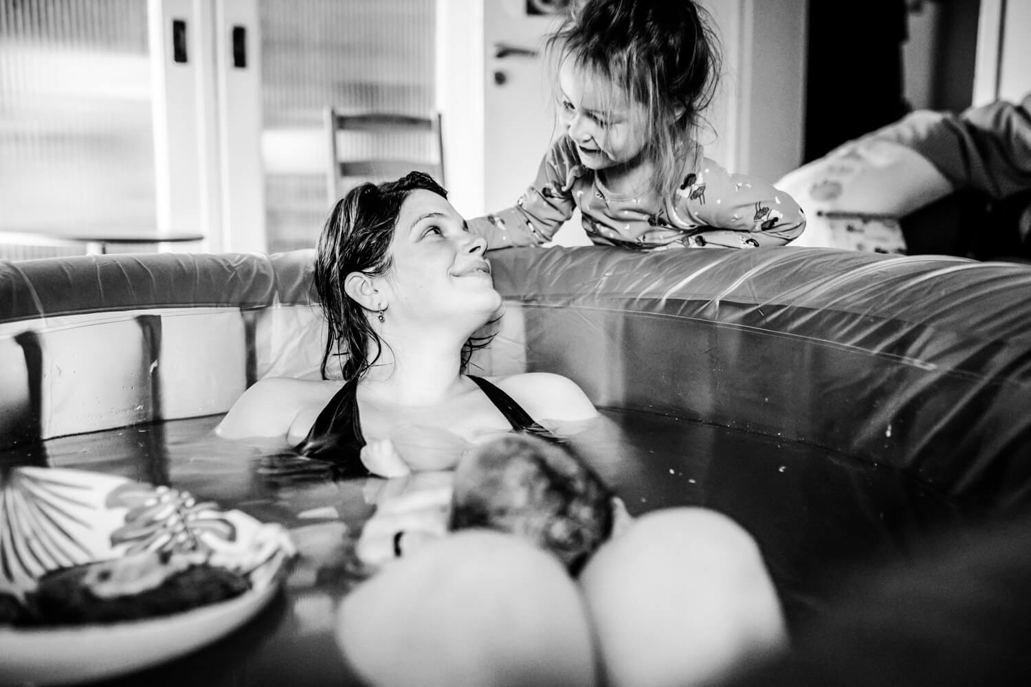 Kleines Mädchen schaut zu Mama, die mit gerade geborenem Baby im Geburtspool sitzt