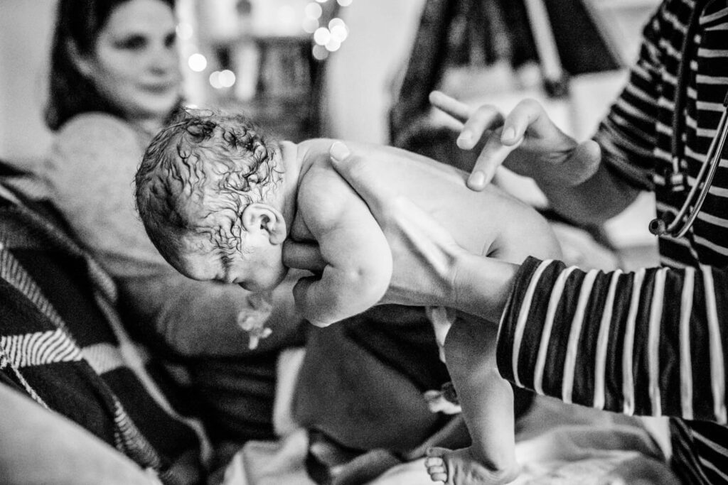 Hebamme untersucht Baby nach Hausgeburt, hält Baby auf ihrer Hand, tastet den Rücken ab
