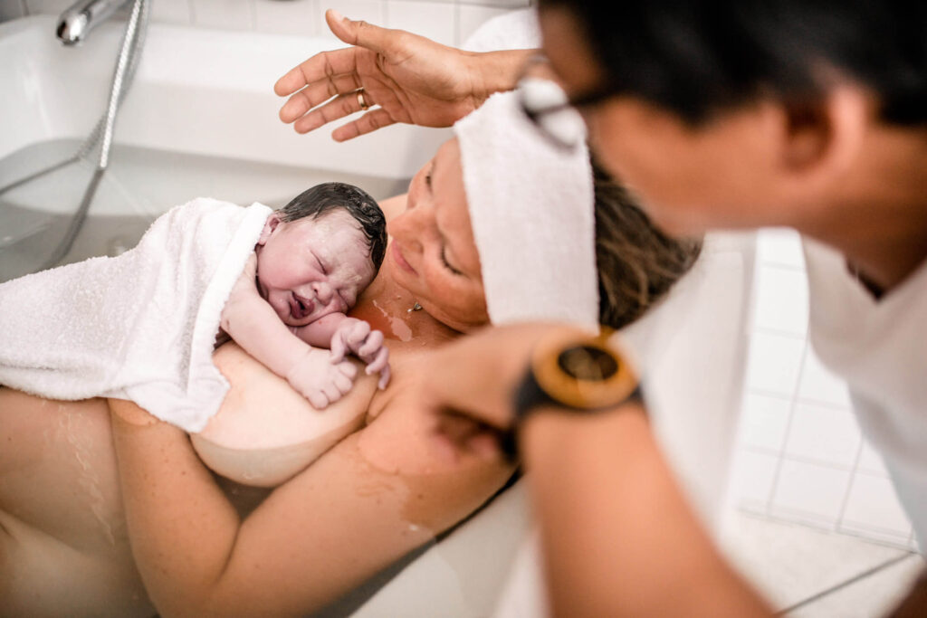 Mutter liegt in Geburtswanne mit gerade geborenem Baby auf dem Arm, Vater sitzt dahinter, Geburtsfotografie Hannover