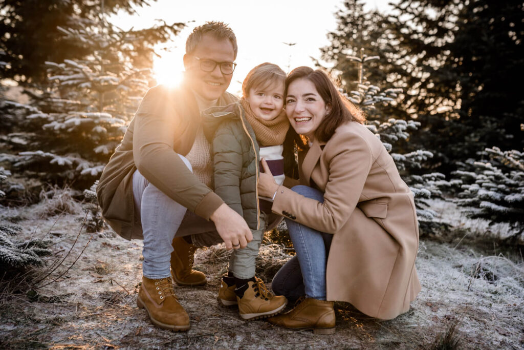 Mutter, Vater, kleiner Sohn hocken vor Weihnachtsbäumen, schauen in die Kamera und lachen, Weihnachtsshooting in Hannover