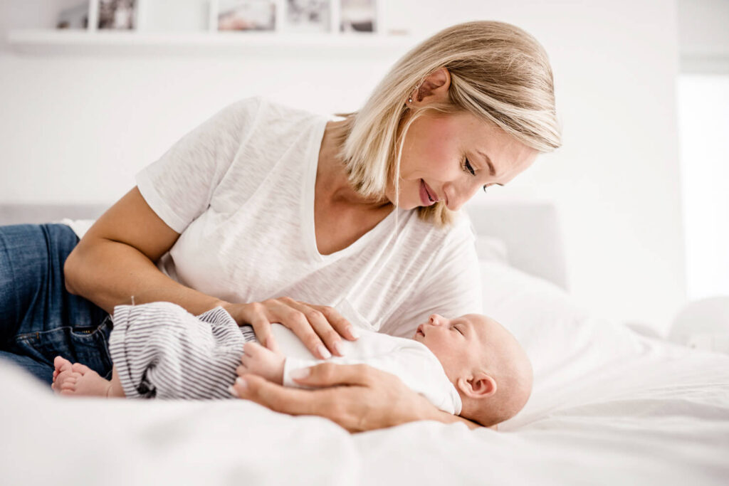 Mutter liegt mit neugeborenem Baby auf dem Bett, schaut schlafendes Baby an, helle Kleidung, weiße Bettwäsche, Fotograf in der Nähe finden