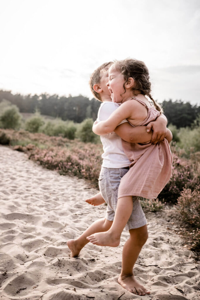 Junge trägt seine kleine Schwester auf dem Arm, sie laufen dabei über den Sand, Fotograf in der Nähe finden