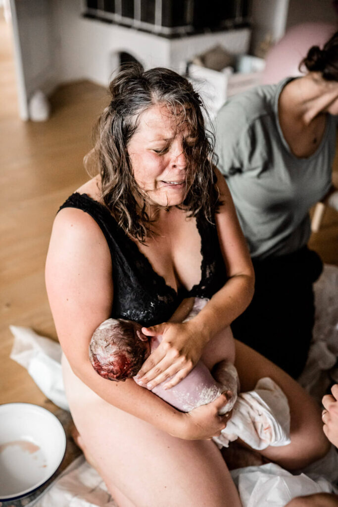Mutter kniet auf Boden und hält gerade geborenes Baby auf dem Arm, Geburtsfotografen in Hannover finden