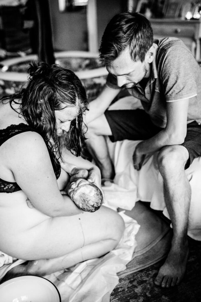 Mutter kniet auf Boden und hält gerade geborenes Baby auf dem Arm, Papa sitzt gegenüber, Geburtsfotografen in Hannover finden