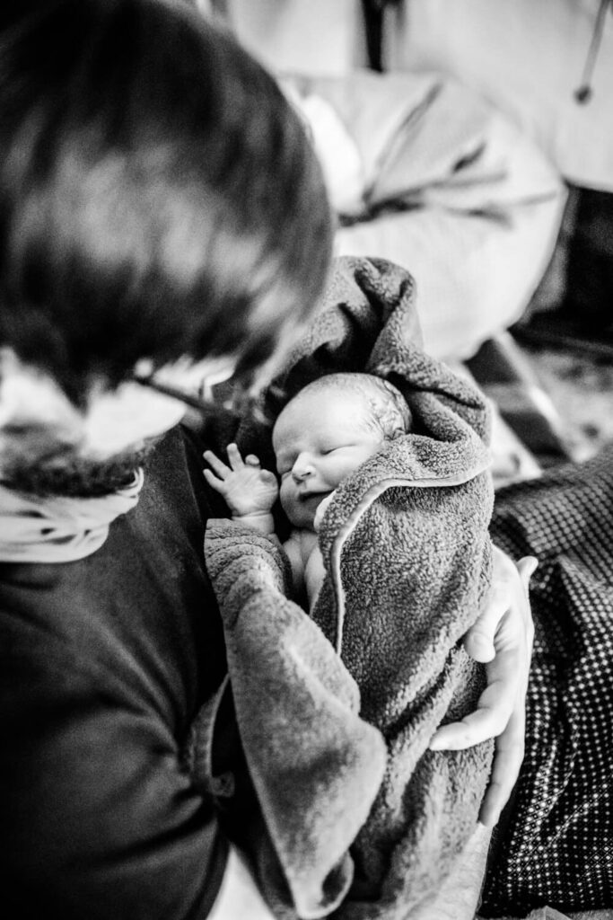 Vater hält gerade geborenes Baby zum ersten Mal im Arm, Baby in Handtuch gewickelt, Geburtsfotos Partner mag nicht