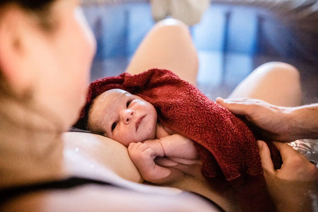 Gerade geborenens baby liegt in rotem Handtuch gewickelt bei Mama auf dem Bauch im Geburtspool, Baby schaut Mama an, wann geburtsfotograf kontaktieren