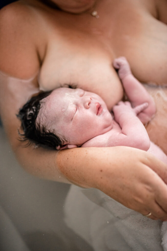 Gerade geborenes Baby liegt bei Mama im Arm in Geburtswanne im Wasser, was ist geburtsfotografie