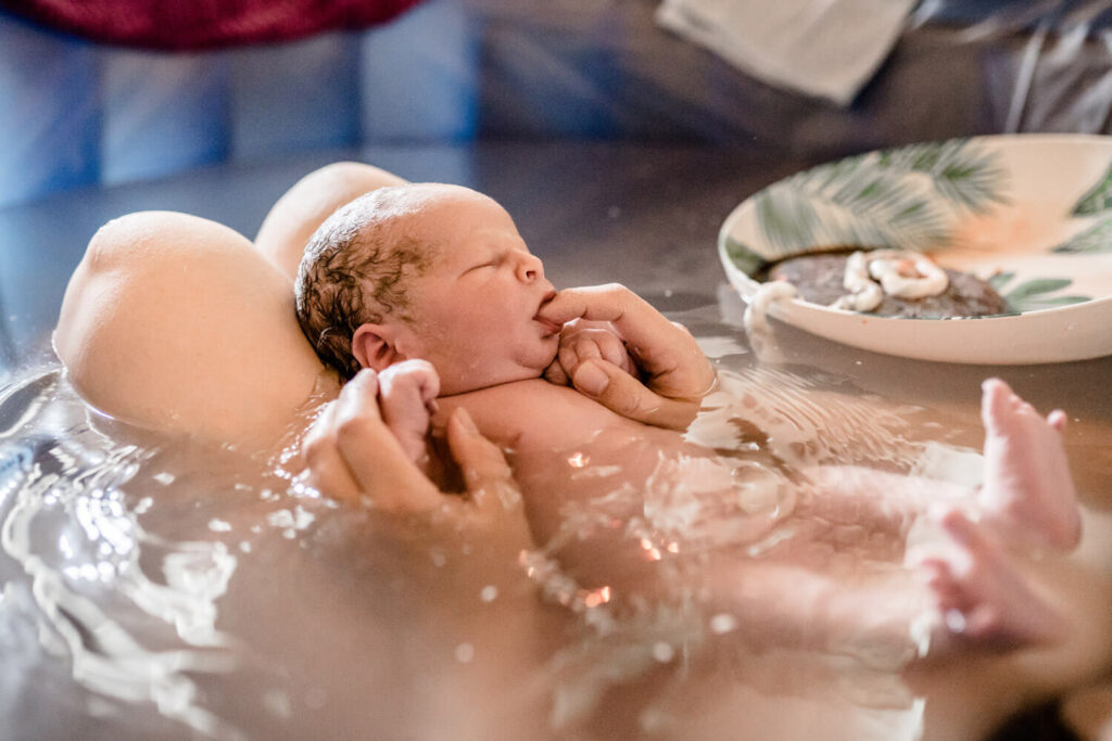 Gerade geborenes Baby liegt auf den Beinen der Mama in einem Geburtspool und saugt an Finger der Mama, was ist Geburtsfotografie