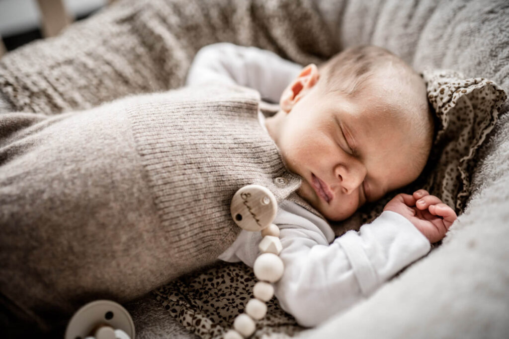 Neugeborenes Baby liegt in Nestchen und schläft, helle, beige Strickkleidung, Schnullerkette aus Holz, Babyshooting mit Julia Schlenkrich