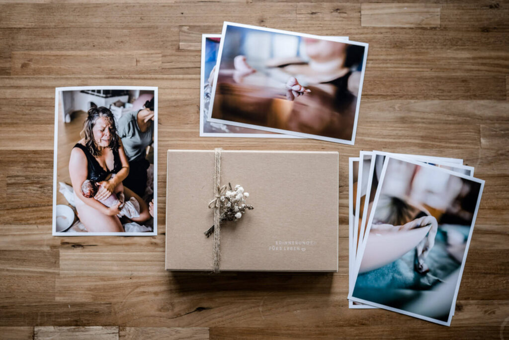 Fotoschachtel aus Kraftpapier liegt auf Holztisch mit Fotoabzügen einer Geburtsreportage, daneben liegt ein Dattezweig, Was kostet ein Geburtsfotograf