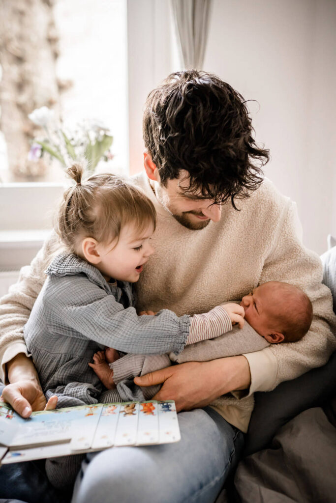 Papa hat Tochter auf dem Schoß und Baby im Arm, lesen ein Bauch und schauen zum Baby, Tochter streichelt Baby während Babyshooting in Hannover