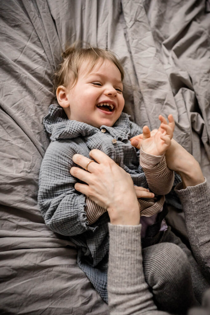 Kleines Mädchen wird von Mama durchgekitzelt, liegt auf Bett und lacht während Babyshooting in Hannover