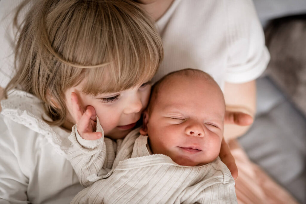 Kleines Mädchen hat neugeborenen Bruder auf dem Arm, kuschelt an seinem Gesicht, Baby hat Augen geschlossen, Babyshooting in Hannover