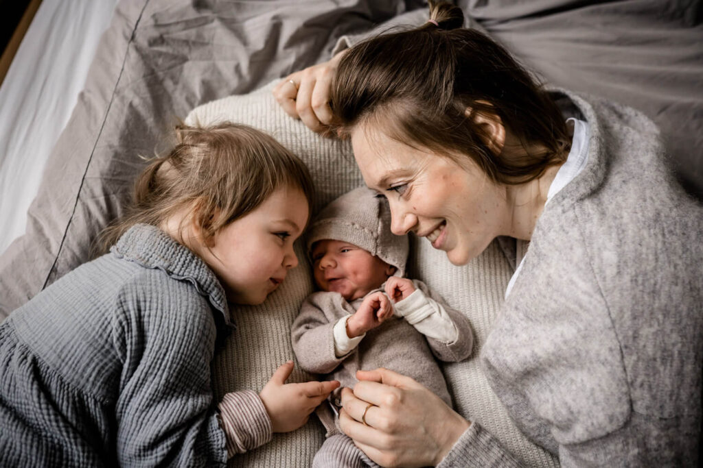 Mutter und Tochter liegen auf Bett, zwischen ihnen liegt neugeborenes Baby, sie schauen es an und lächeln, wohnung ordentlich Newborn Shooting