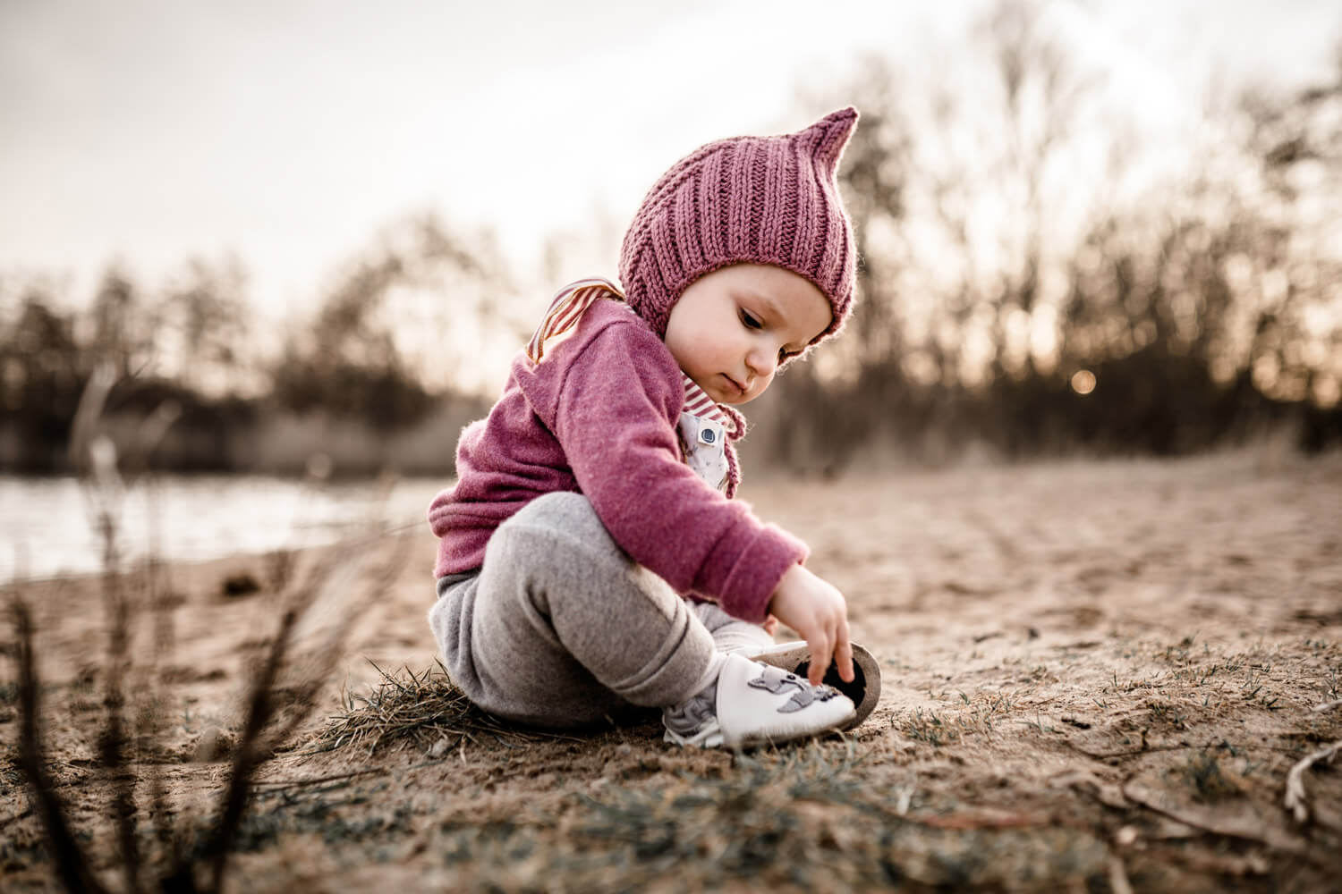 Kleines Mädchen mit pinkter Strickmütze sitzt auf Sand am See und schaut zu ihren Schühchen, Familienshooting im Frühling in Isernhagen, Familienshooting wann machen